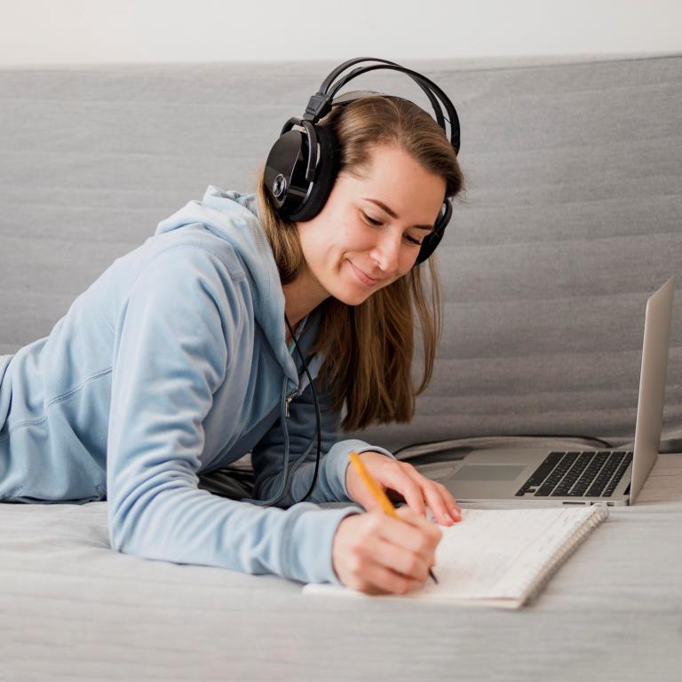 Student wearing headphones and taking notes while taking online classes on her laptop