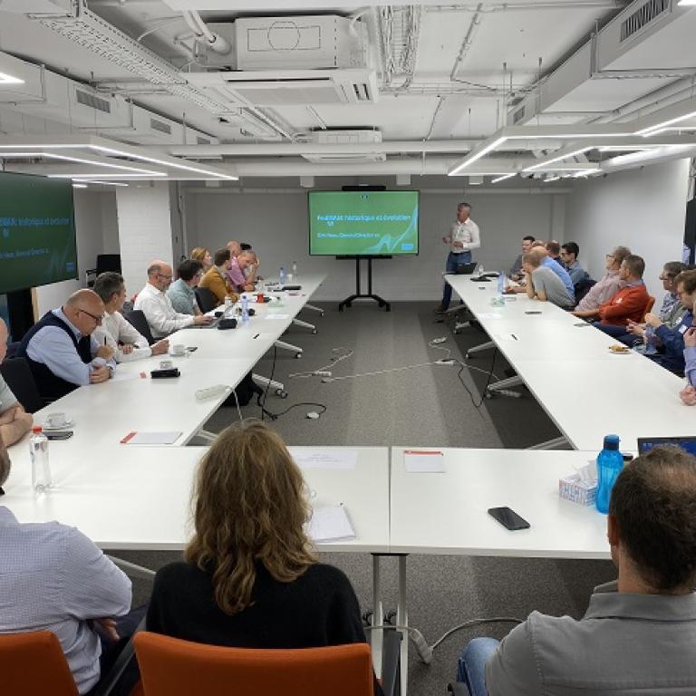 Photo of the meeting room with participants seated around a U-shaped table. The speaker stands near the screen.